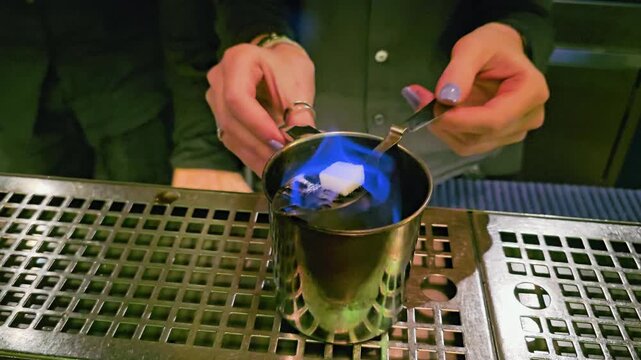 Traditional European absinthe service with flaming sugar cube on a slotted spoon, metal cup, clear goblet, bartender hands, green reflections, and moody bar counter close-up.
