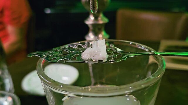 Traditional absinthe louche preparation with sugar cube, ornate slotted spoon, dripping fountain water, cloudy pale cocktail, dark wood table, and green backlit bar ambience.