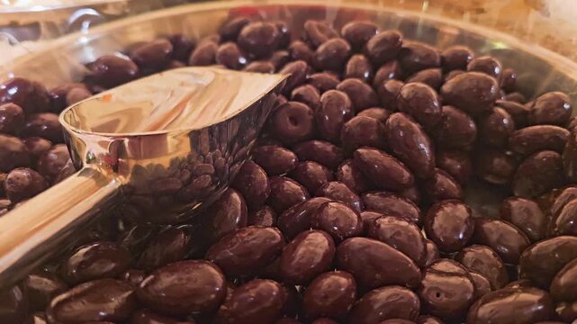 Detailed snack counter shot of chocolate coated nuts piled high in a clear tray, with serving scoop reflections, warm retail lighting, and tempting bulk candy shop texture.