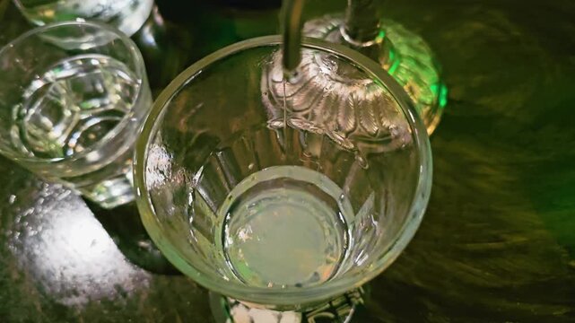 Overhead view of chilled water dripping into absinthe, creating a cloudy louche effect in a cocktail glass beside water glass and reflections on a dark wooden bar table.