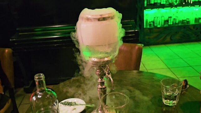 Close-up of dramatic absinthe service with louche fountain pouring into a glass while fog rolls over a round bar table, liquor bottle, spoon, and dim green cocktail lounge lighting at night.