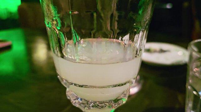 Macro close-up of absinthe louche swirling in a glass, showing milky pale drink, cut crystal texture, green bar reflections, shallow focus, and moody cocktail lounge atmosphere at night.