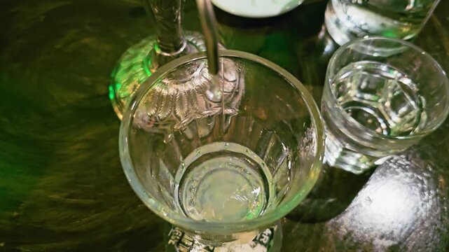Top-down close-up of absinthe louche forming in a glass as cold water drips from a fountain spout, with cloudy pale liquid, reflective bar table, and moody green lounge lighting.