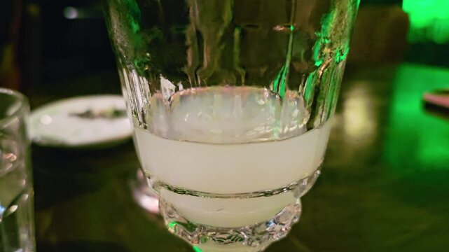 Extreme close-up of cloudy absinthe louche inside a cocktail glass, with creamy white liquid, textured glass details, dark table reflections, and green nightlife lighting in a dim bar.