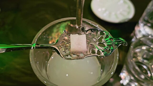 Detailed view of classic absinthe preparation with sugar cube, perforated spoon, dripping water, milky glass, liquor bottle, dark wooden table, and soft green nightlife lighting.