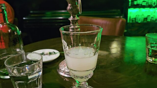 Absinthe louche forms in a vintage goblet under a dripping fountain spout, with cloudy pale drink, bottle, water glass, dark wooden table, and dim green cocktail lounge atmosphere.