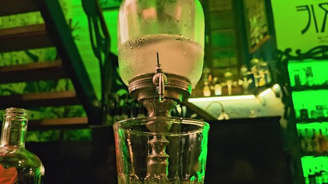 Detailed view of an absinthe louche fountain above a cocktail glass, with chilled water, vintage metal spout, green bar lighting, bottle reflections, and dark lounge interior at night.