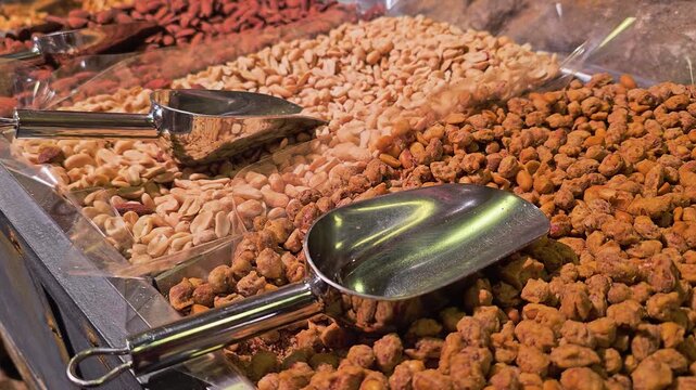 Detailed view of assorted nuts in clear bulk bins, showing cashews, peanuts, almonds, and coated snack mix with metal scoops, warm indoor light, and self serve market display.