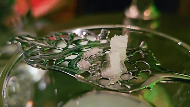 Extreme close-up of absinthe service as water melts a sugar cube on a slotted spoon, showing sparkling crystals, foamy texture, glass rim, and moody green lounge lighting.