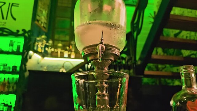 Close-up of absinthe louche dripping from an ornate fountain into a glass, with green backlit bottles, staircase, liquor bottle, and moody cocktail bar atmosphere indoors at night.