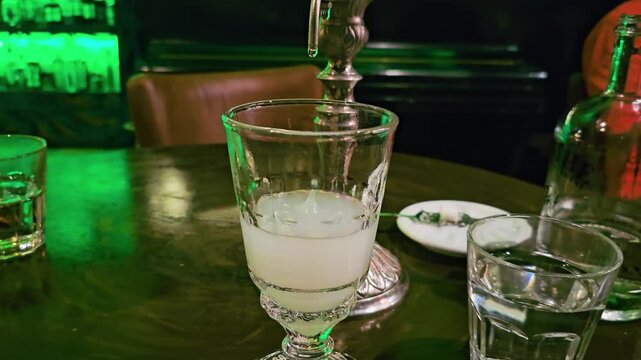 Close-up of absinthe louche in a glass as water drips from an ornate fountain, with milky cocktail, water glass, liquor bottle, spoon, green bar lighting, and moody nightlife table scene.