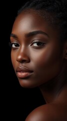 Fototapeta premium A close-up portrait of a dark-skinned woman with smooth skin, expressive eyes, and natural makeup against a black background.