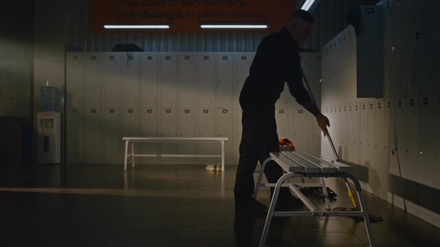 Tattooed Male Custodian Working inside Spacious Sports Locker Room. Man Systematically Cleans Premises, Mops Under Benches Where Soccer Ball and Sports Bag Left, and Closes Open Metal Cabinets.
