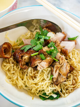Dry Egg Noodles with Roasted Duck and Crispy Pork Belly Topped with Fresh Herbs, Served with Clear Soup on Marble Table