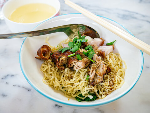 Dry Egg Noodles with Roasted Duck and Crispy Pork Belly Topped with Fresh Herbs, Served with Clear Soup on Marble Table