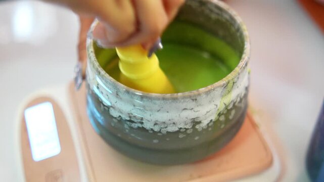 4K Close up of hand using traditional whisk to mix green matcha tea powder in clear glass bowl, Concept of traditional Japanese tea ceremony preparation and healthy lifestyle.