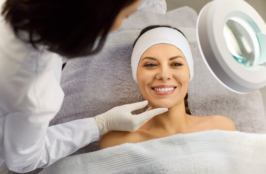 Professional female beautician or cosmetologist examining skin condition to a young smiling pretty girl client with special lamp in beauty spa salon. Skin care and treatment concept.