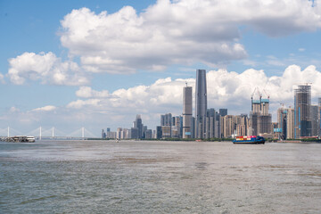 The modern city skyline of Wuhan China stands tall along the river with cargo ships passing by on a bright sunny day under a beautiful cloudy sky © AS_SleepingPanda