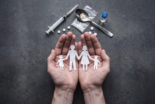 Protective hands holding a paper cut-out family surrounded by drug paraphernalia, symbolizing the impact of addiction on family life and safety.