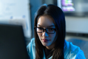 Fototapeta premium Close-up of an Asian woman in glasses looking down at her screen with focus, lit by the cool blue glow of a dark programming office at night.