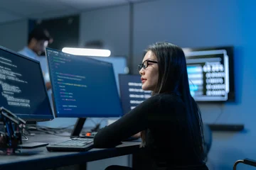 Asian female developer in glasses works at a row of monitors filled with code and data while a colleague works in the background of a dark office. © Jestercine