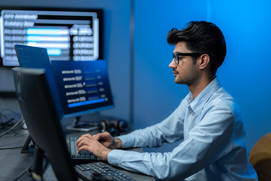 South Asian man in glasses types at his keyboard with a data dashboard glowing behind him against a deep blue office wall at night.