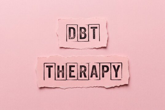Dialectical Behavior Therapy concept with the letters DBT and THERAPY printed on textured pink paper scraps against a soft pink background.