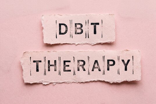 Dialectical Behavior Therapy (DBT) spelled out with stamped letters on torn paper strips against a soft pink background.