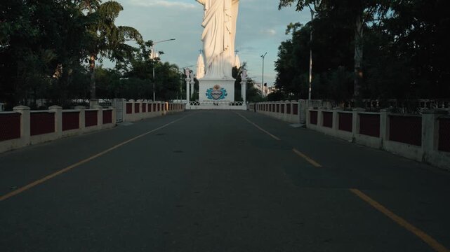 Cinematic gimbal push-in shot along a tree-lined road leading to the towering Sacred Heart of Jesus statue in Velankanni, arms raised against a soft morning sky in Tamil Nadu, India.