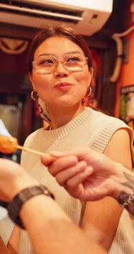 Man, feeding and asian woman with skewer for japanese cuisine, travel or lunch together. Female person, stick or eating with kebab, dango or street food for meal, snack or dining in town market