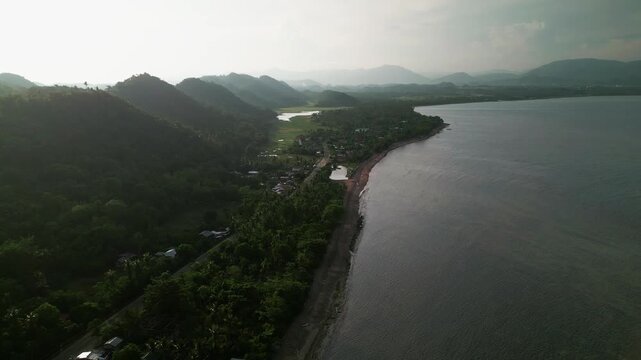 Aerial view of lush mountains, tropical coastline, and a narrow coastal village stretching along shore - Catanduanes, Bicol, Philippines