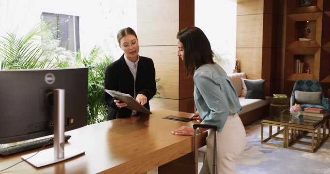 Young woman receptionist holds clipboard showing or explaining guest registration form to female, client stands at counter receive information about hotel services or list of amenities. Accommodation