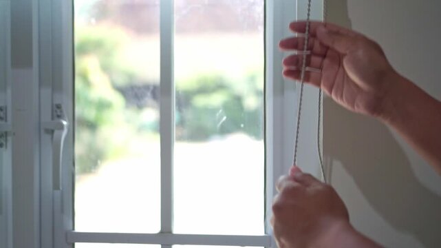 Hand of person opening a window curtain by pulling the cord. Bright daylight fills the room, showcasing comfort, home design, and everyday routine.