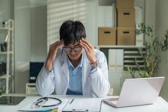 Stressed young doctor wearing glasses holding his head with hands showing fatigue and headache at clinic desk.