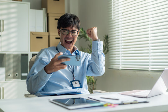 Excited young businessman raising fist celebrating success while looking at smartphone at office desk.