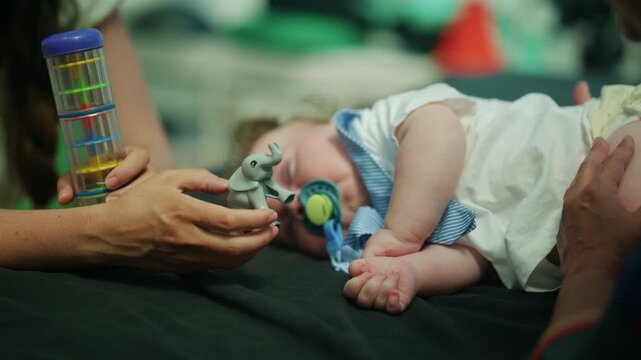 Parent carefully holding a rattle and an elephant toy while a baby is sleeping peacefully on a dark surface, wearing a pacifier