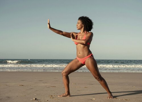 Woman practicing martial arts pose barefoot on sandy beach ocean waves background vibrant coral bikini athletic movement