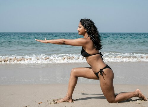 Woman practicing yoga pose on beach during sunrise; black bikini, ocean waves, serene motion