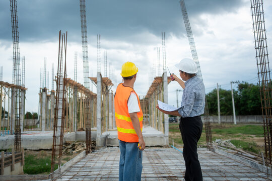 Two engineers in hard hats review plans at a building site with steel columns under dark storm clouds, showing teamwork, planning, safety and construction progress.