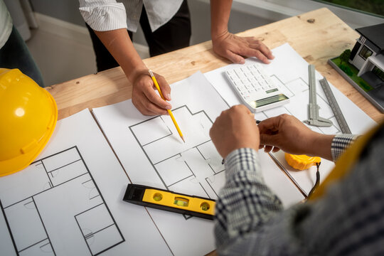 Architects and builders review house plans on a wooden table with tools, calculator and hard hat, showing teamwork, planning and construction design.