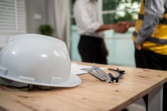 White safety helmet on a wooden desk with work tools, while two people shake hands in the background, showing agreement, construction planning, and workplace safety indoors.