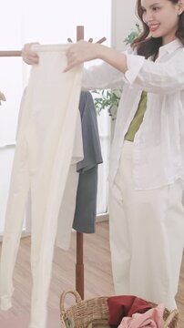 A smiling Asian woman doing laundry at home. She hangs a t-shirt on a wooden clothes rack.