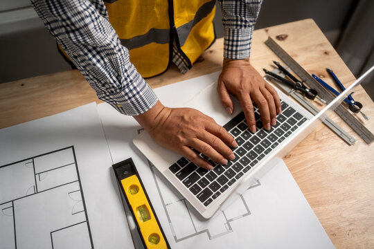 Engineer or builder using a laptop over house plans at a work desk with measuring tools and safety vest, showing planning, design, and construction work.