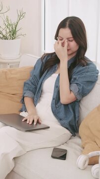 Tired young Asian woman closing her laptop, rubbing her eyes, and stretching after working from home.