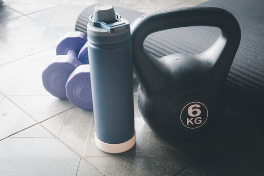 Close up of home workout and gym equipment featuring a black 6kg kettlebell, blue water bottle, purple dumbbells,and a rolled yoga mat on a tiled floor.Strength training and active lifestyle concept.