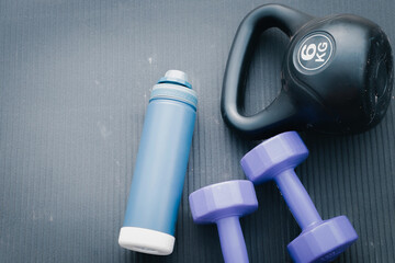 Fototapeta premium Top view flat lay of fitness equipment featuring a blue water bottle, two purple dumbbells, and a black 6kg kettlebell on a grey textured gym mat with copy space for text.