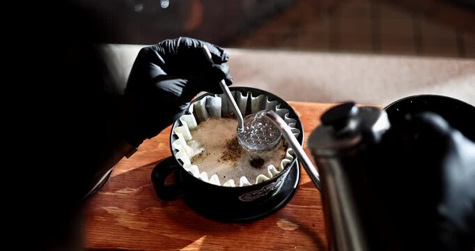 Gloved hand stirs blooming pour over coffee while hot water flows from a gooseneck kettle indoors