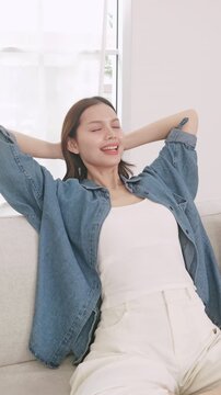 A relaxed young Asian woman sitting on a sofa, stretching her arms and resting with her eyes closed at home.