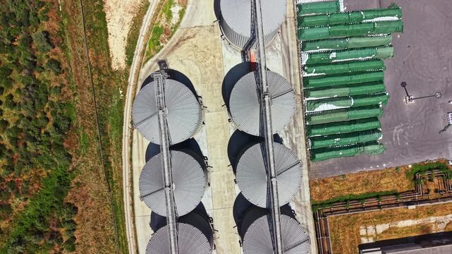 Aerial view shows large storage silos arranged in rows next to green containers in an industrial site. There are fields and trees in the background. This scene captures an active area.