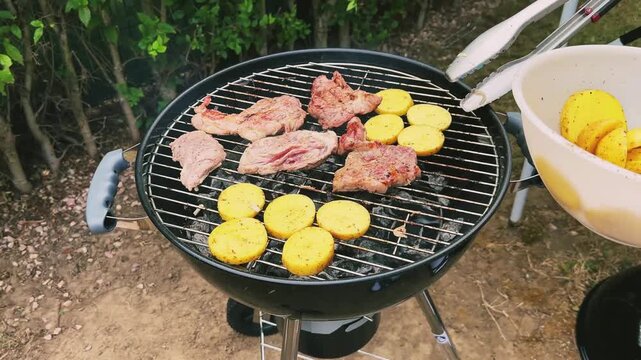 Meat and potatoes are grilled over a fire. The cook puts fresh potatoes on the grill with tongs. Backyard BBQ. High quality 4k footage